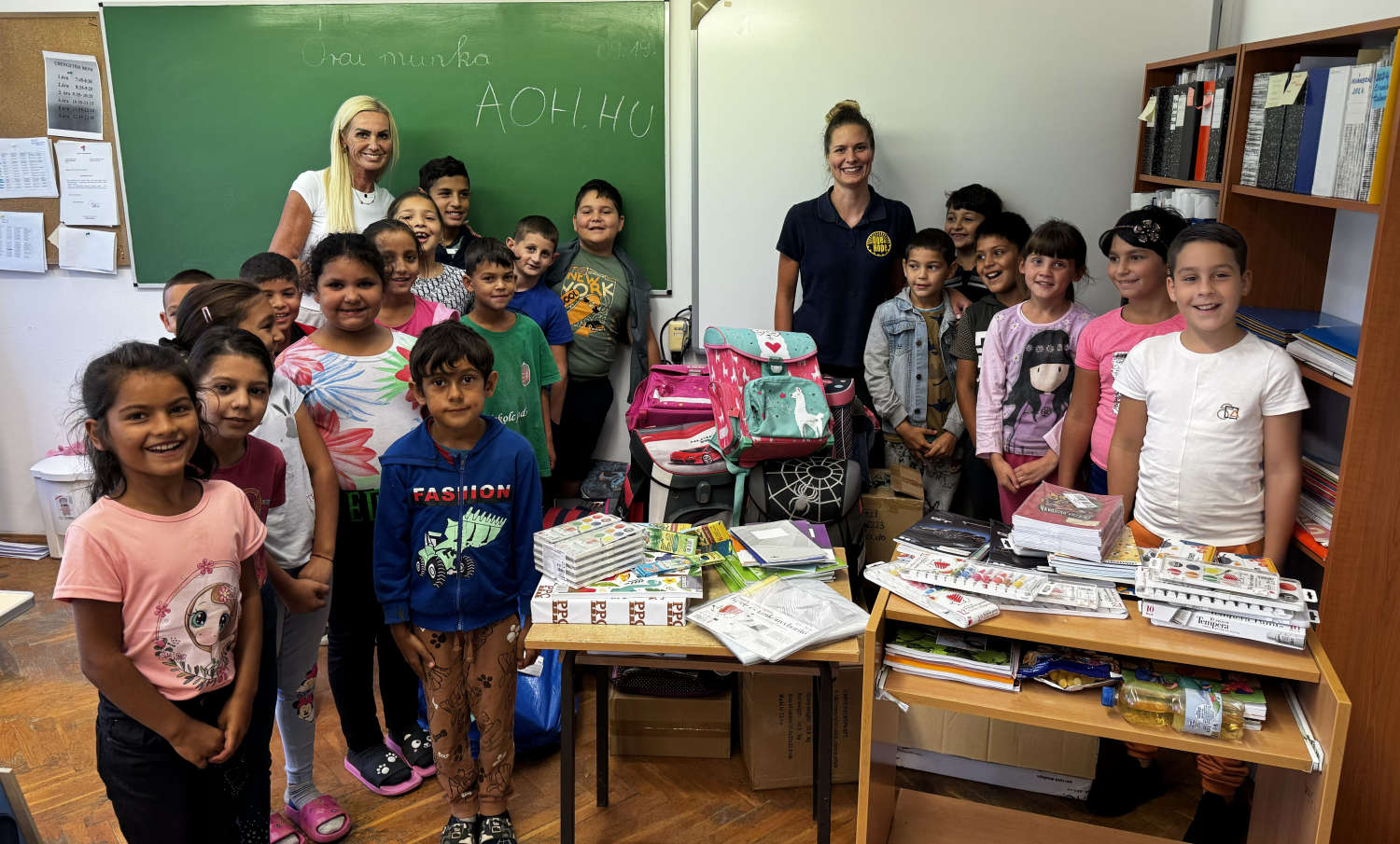 Smiling children in a classroom with new backpacks and school supplies, AOH written on the board