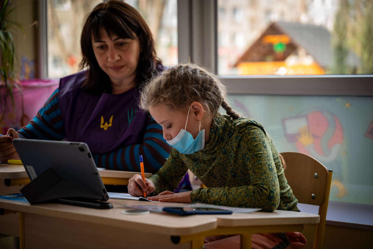 Teacher helping a masked girl learn on a tablet at school